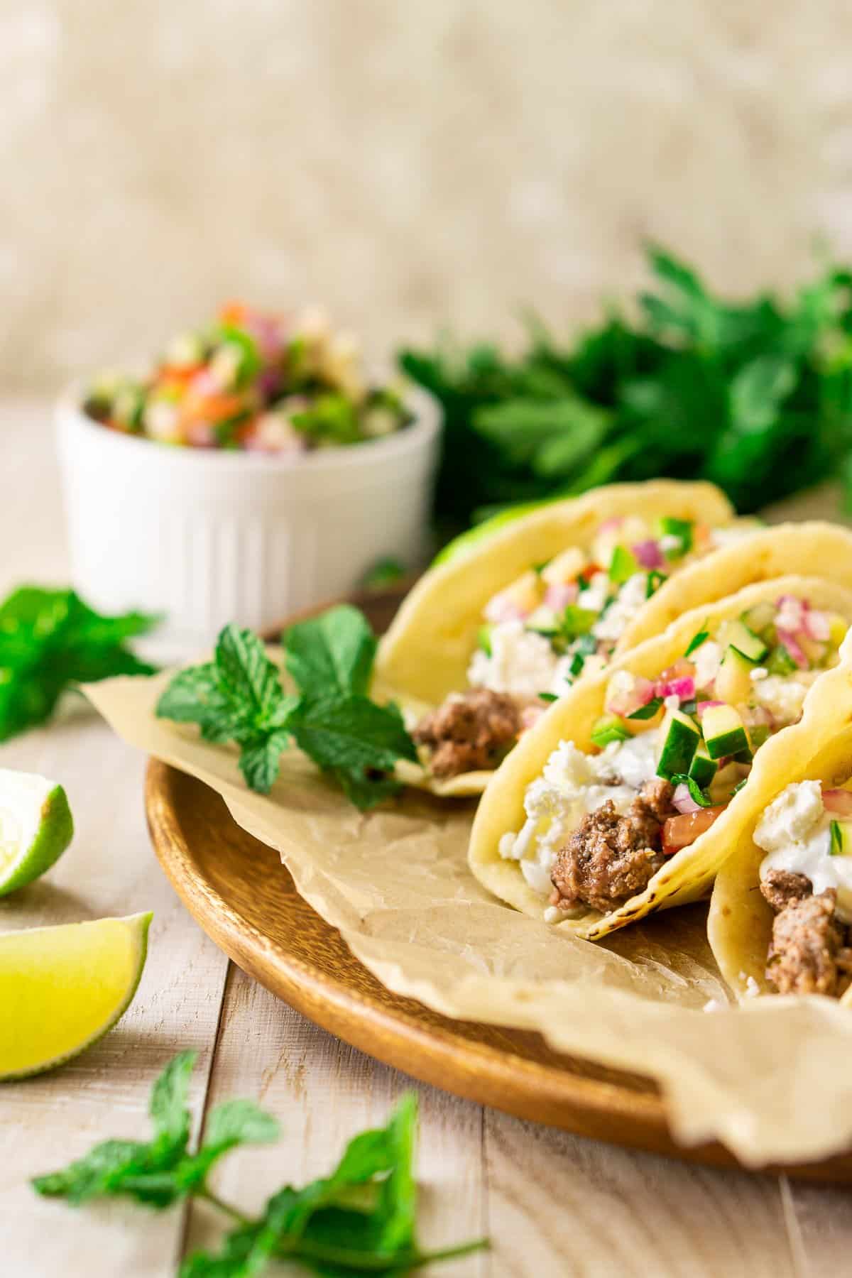 Greek lamb tacos with cucumber pico de gallo on a parchment paper lined wooden platter with more pico in a serving dish.