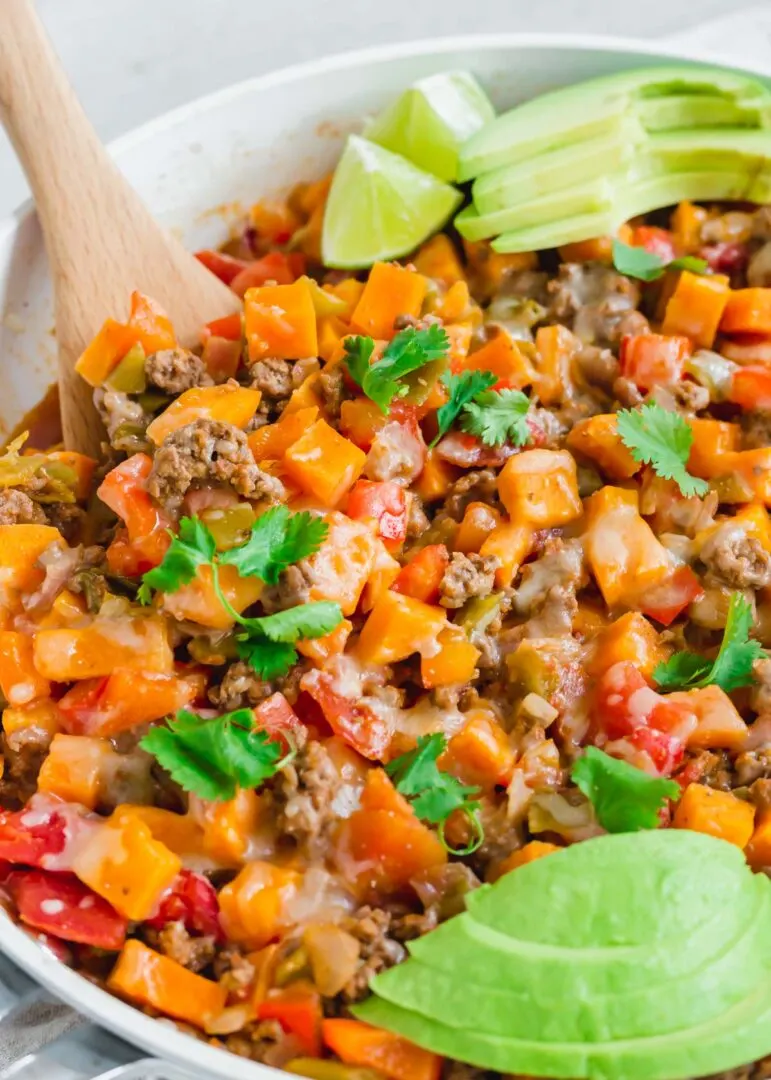 Ground beef and sweet potato skillet with avocado in a casserole dish with wooden spoon.