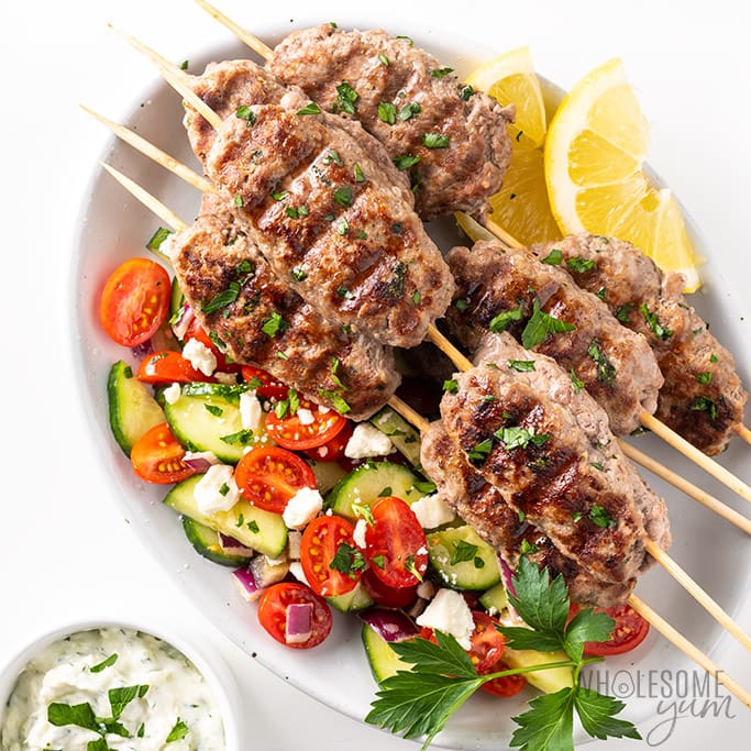 Grilled ground lamb with Mediterranean salad and lemon on a white platter.