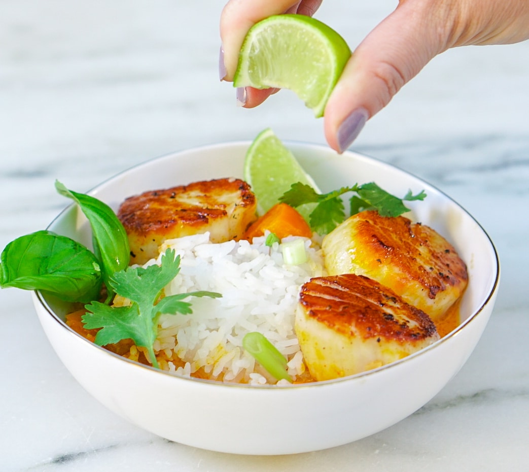 Hand squeezing lime over bowl filled with curry scallops, and rice.