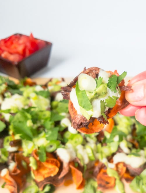 Fingers holding sweet potato nachos with shredded beef and toppings.