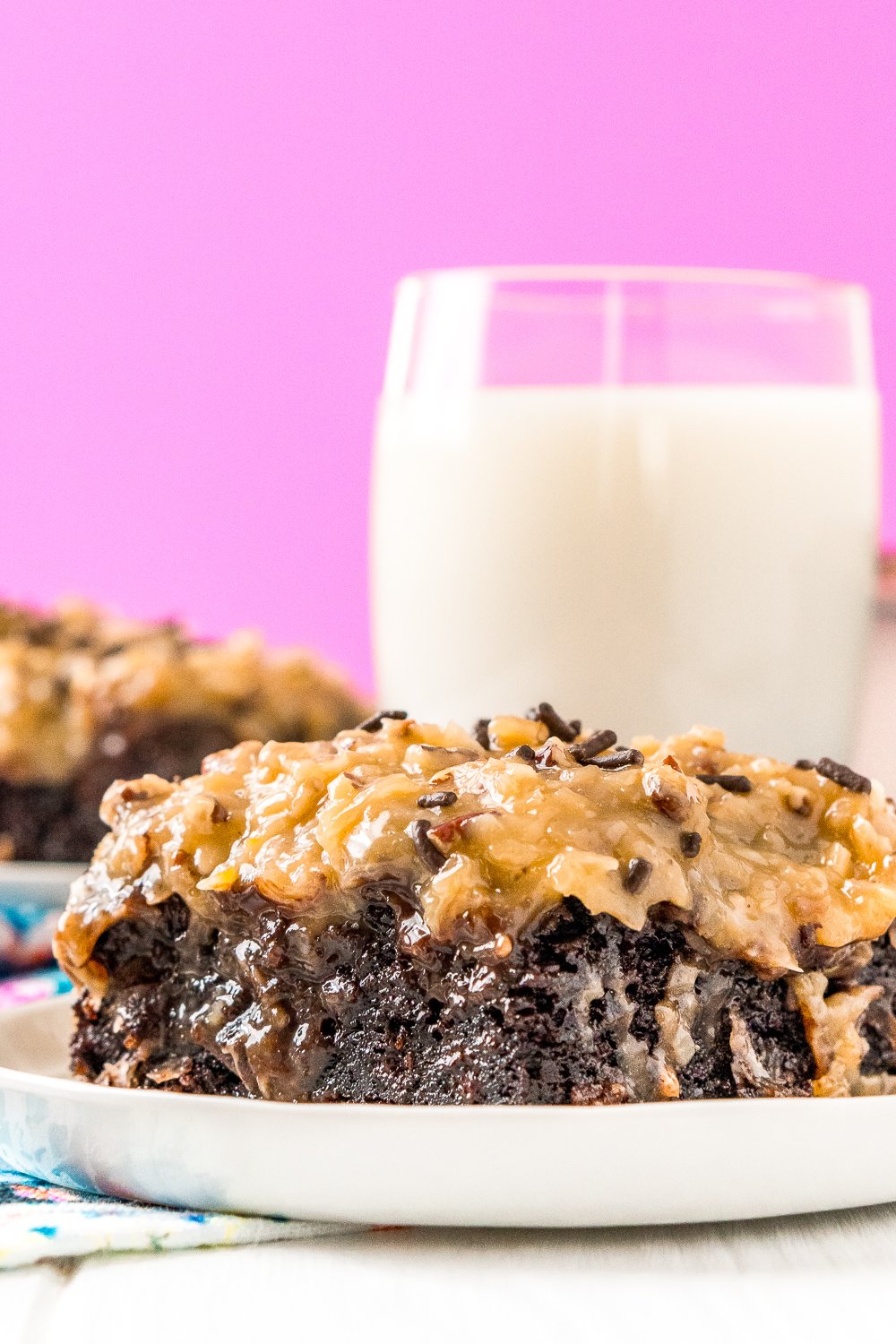 Slice of moist German chocolate cake on a white plate with a glass of milk in the background.