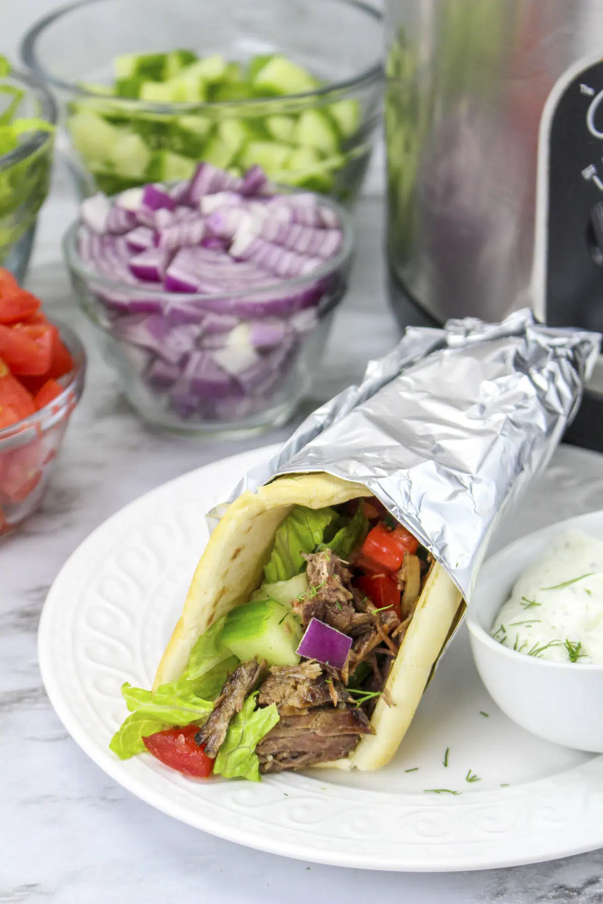 Slow cooker beef gyro on a white plate with tzatziki sauce with ingredients in a clear glass bowls.