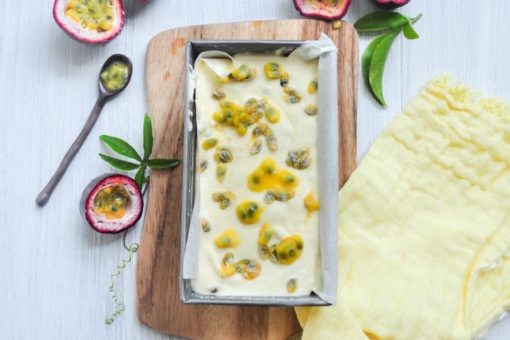 Passionfruit ice cream in a container with spoon and passionfruit in background.