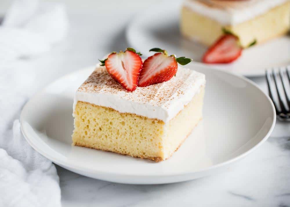 Slice of tres leches cake topped with slices of strawberry on a white plate.