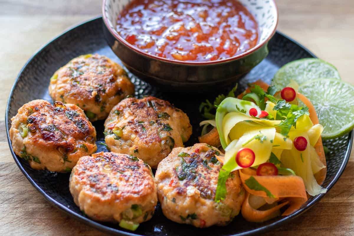 Thai fish cakes on a plate with dipping sauce and vegetables.