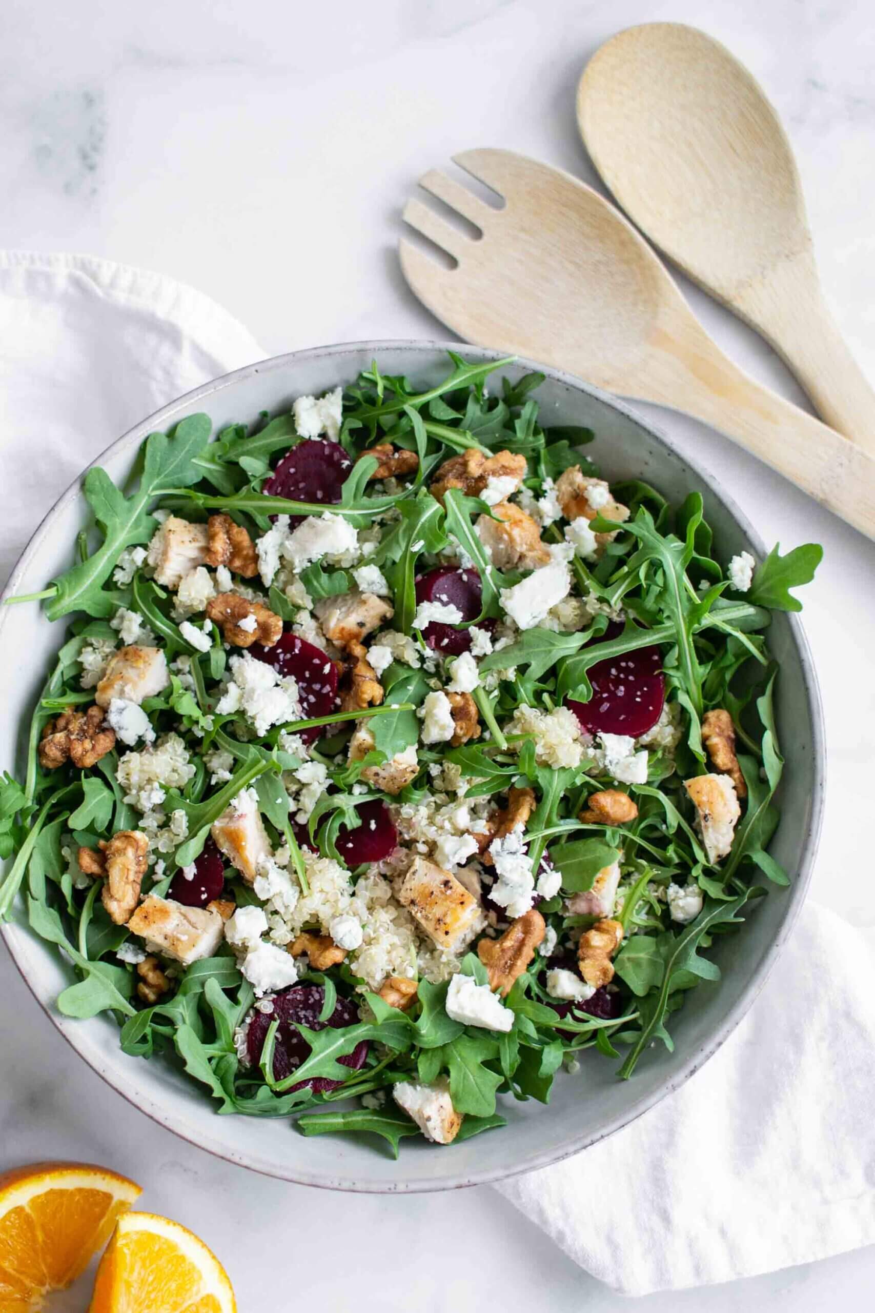 Salad with quinoa, diced chicken feta, beets, and arugula in a bowl with soon and fork, sliced oranges in the background.