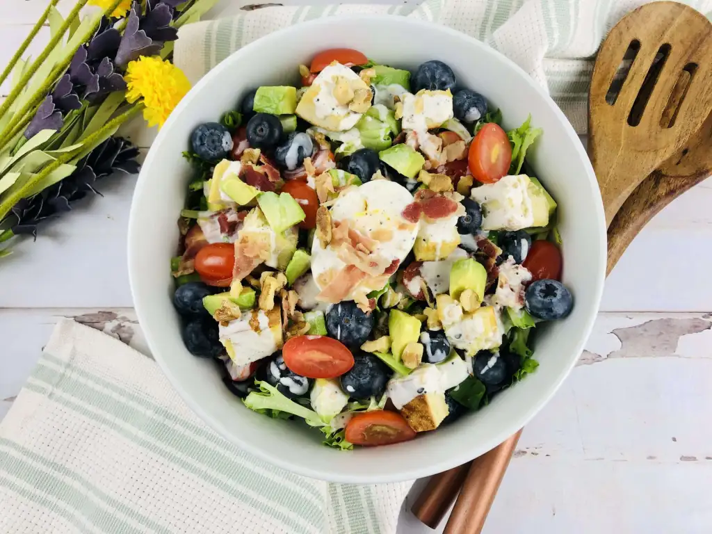 Salad with avocado, tomatoes, blueberries, eggs, and diced chicken with flowers, wooden salad utensils, and napkins on a white background.