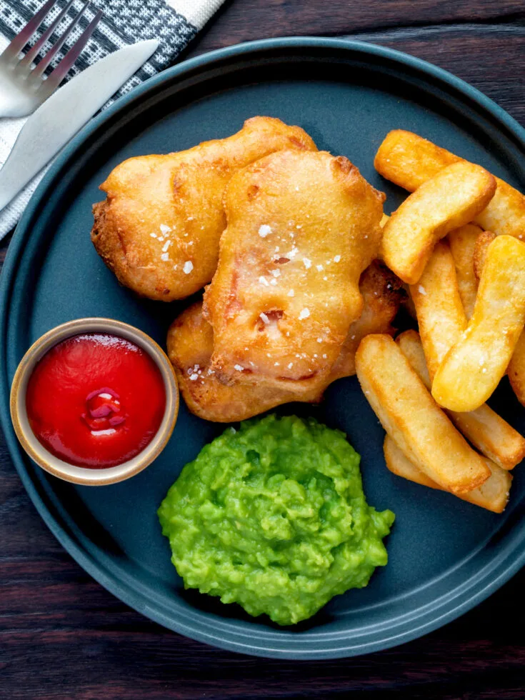 Beer battered spam fritters with fries, ketchup, and mushed peas on a blue plate.