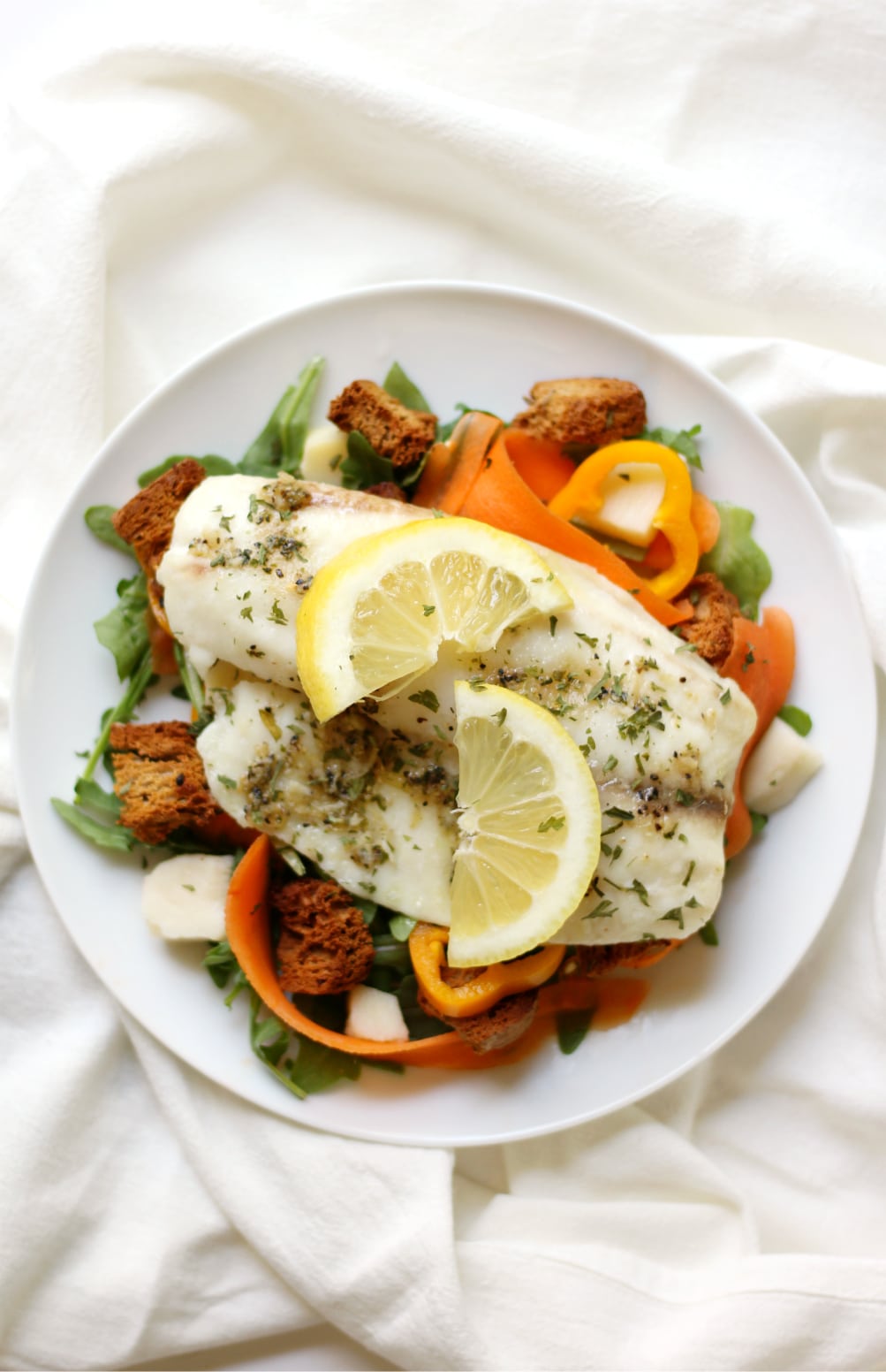 Lemon pepper seasoned tilapia on a plate with arugula, bell pepper, shaved carrots, and croutons on a white plate.