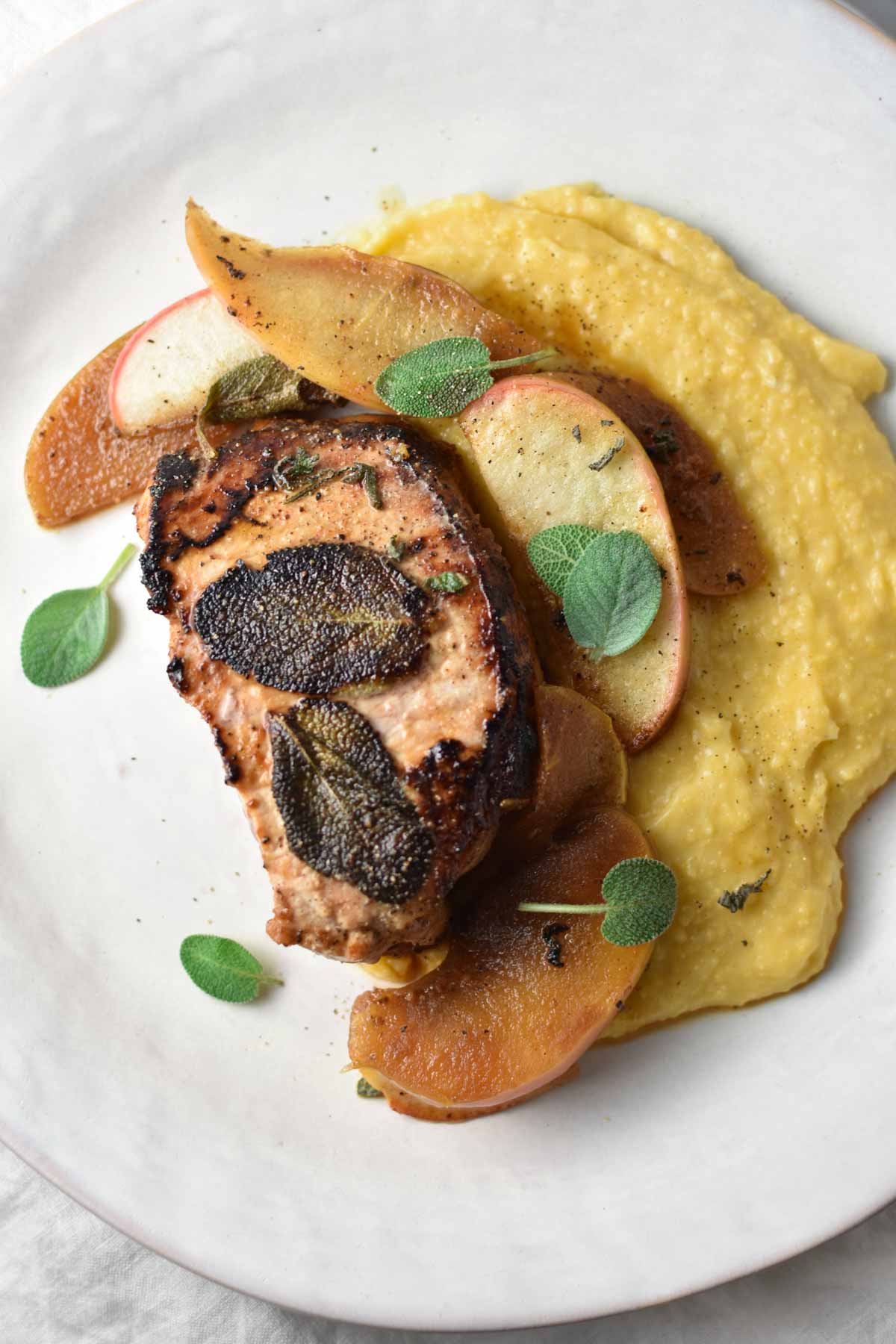 Pork chops with apple and sage on a white plate with polenta.