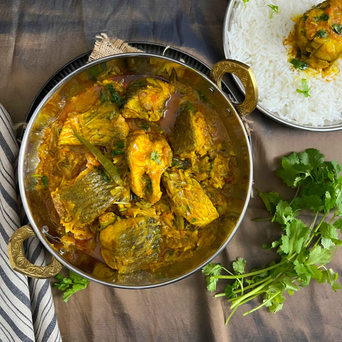 Bengali fish curry in a pot with rice and cilantro in the background.