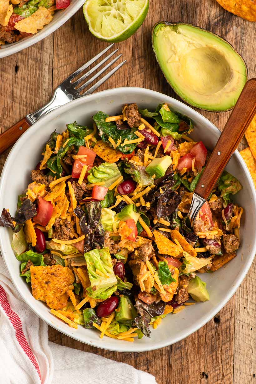 Dorito taco salad recipe in a bowl with ground beef, avocado, lettuce, tomatoes, cheese, and crushed Doritos with a fork in the background.