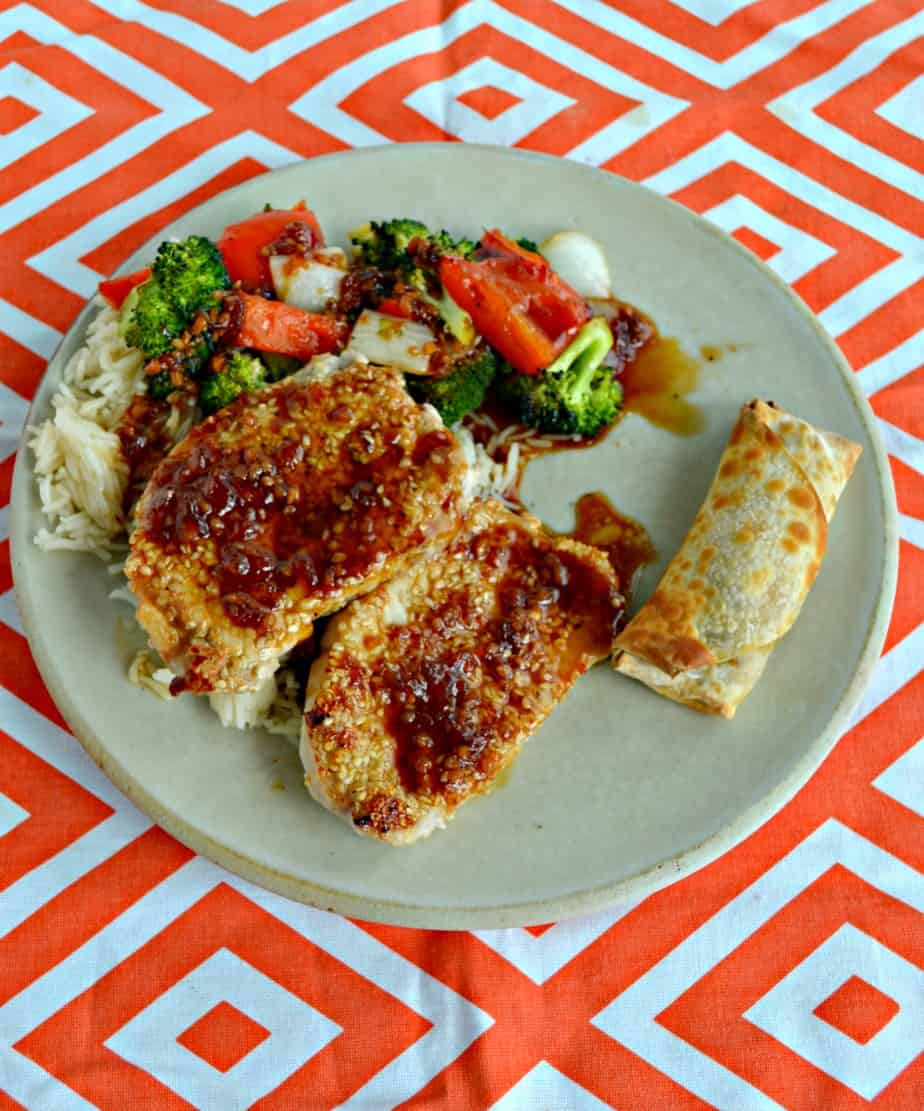 Sesame crusted pork chops on a plate with vegetables, rice, and an egg roll.