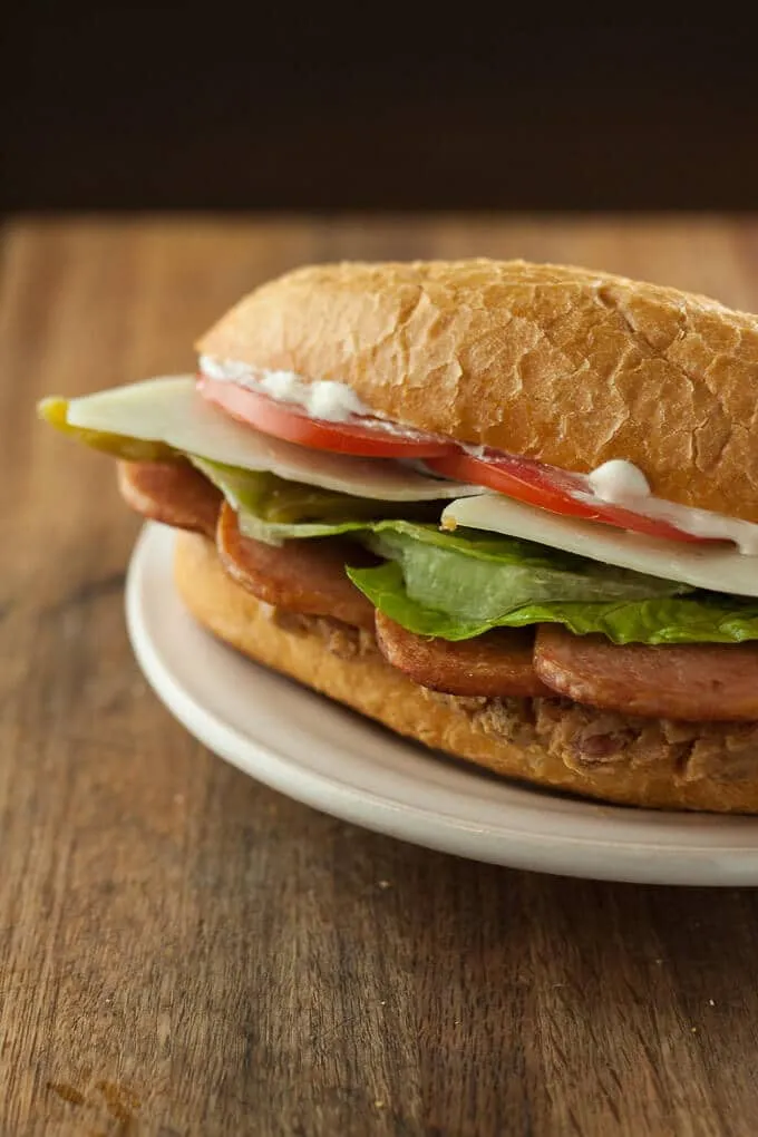 Spam sandwich with lettuce cheese tomatoes and mayo on a white plate.