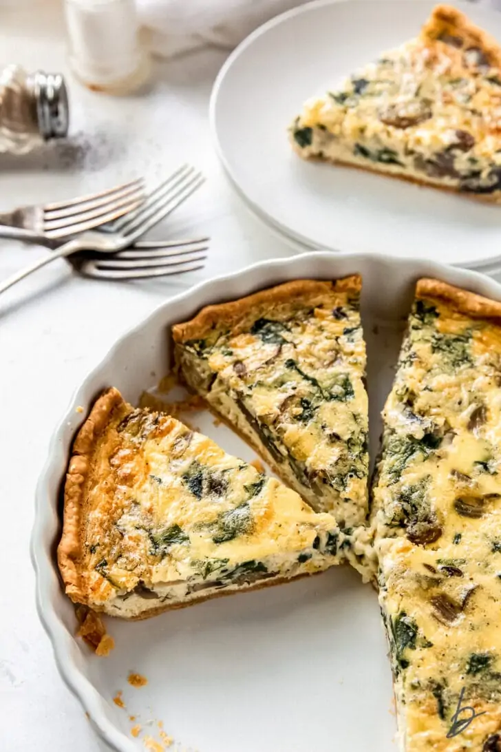 Spinach mushroom quiche in a baking dish and a slice on a plate in the background.