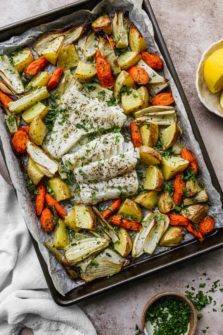 Herb flavored cod and vegetables in a sheet pan with lemon on a separate dish.