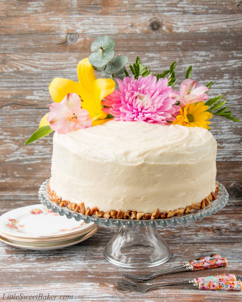 Hummingbird cake topped with flowers on a clear cake stand with decorative plates and forks on a wooden background.