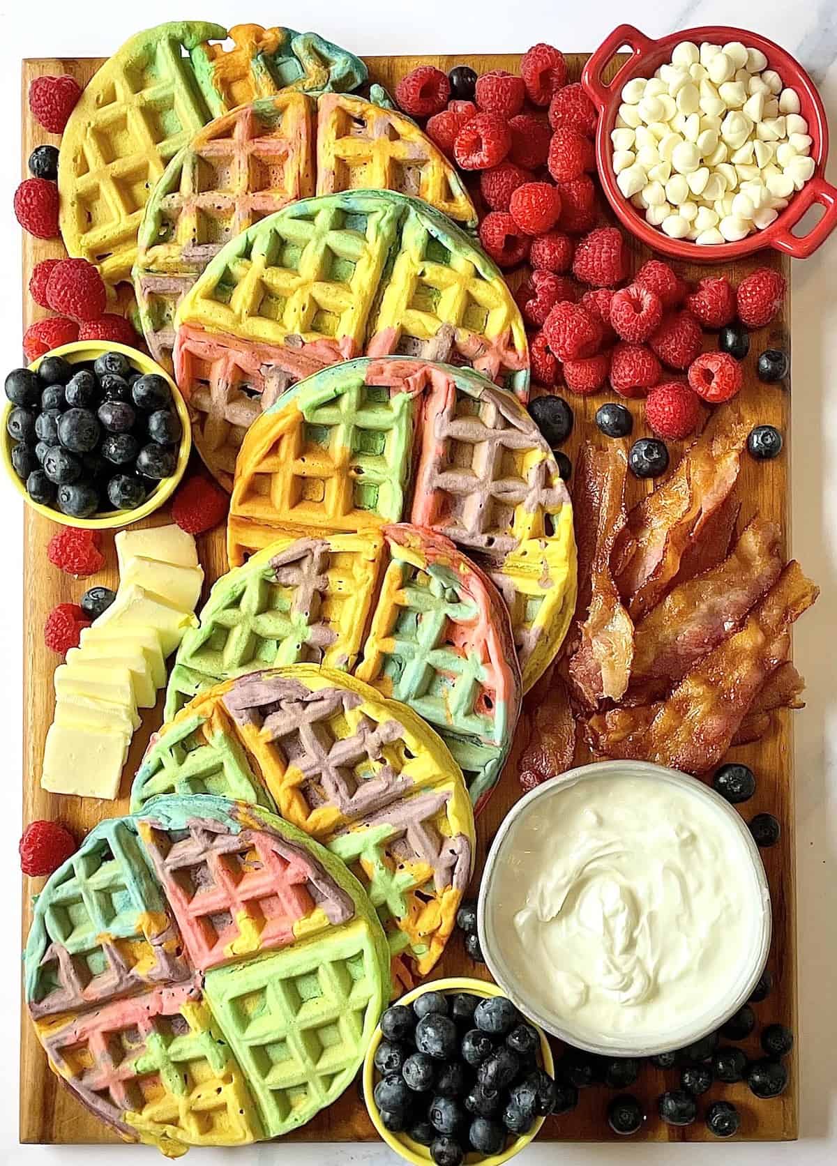 Rainbow waffles charcuterie board with with berries, white chocolate chips, butter, and whipped cream.