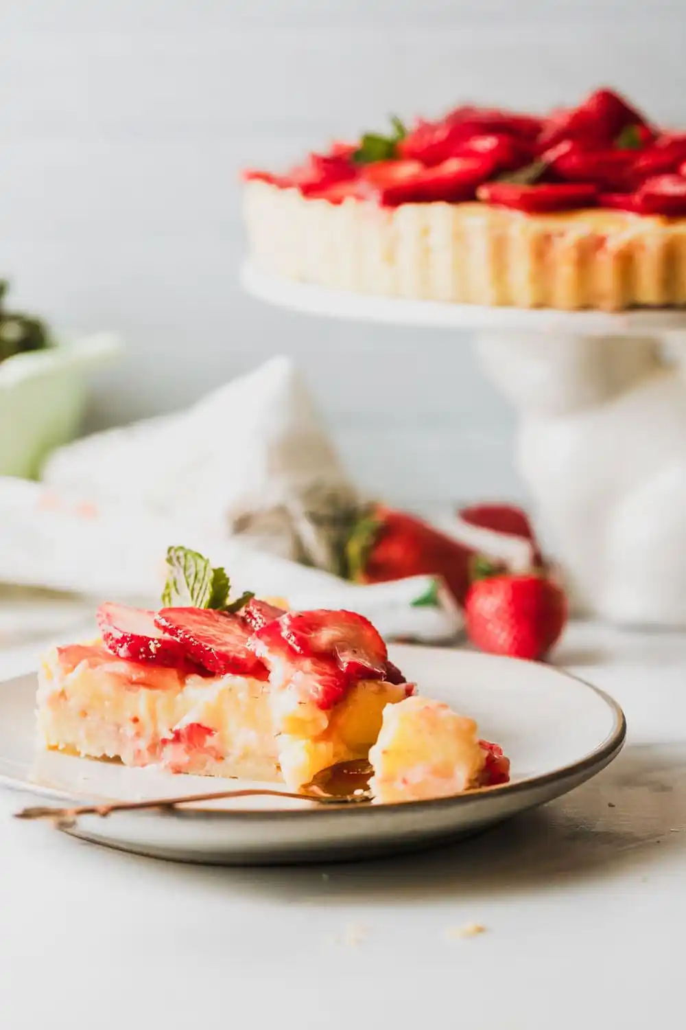 Slice of strawberry custard tart on white plate with rest of tart in the background.