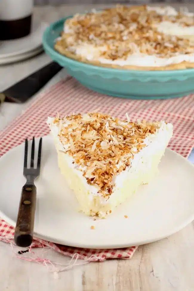 Slice of coconut cream pie on a white saucer with more in background.