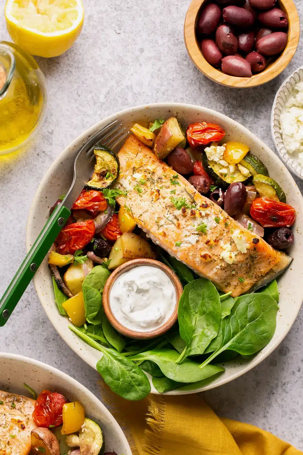 Sheet pan Greek salmon with olives, tomatoes, potatoes, onions, and spinach with a fork in a bowl.