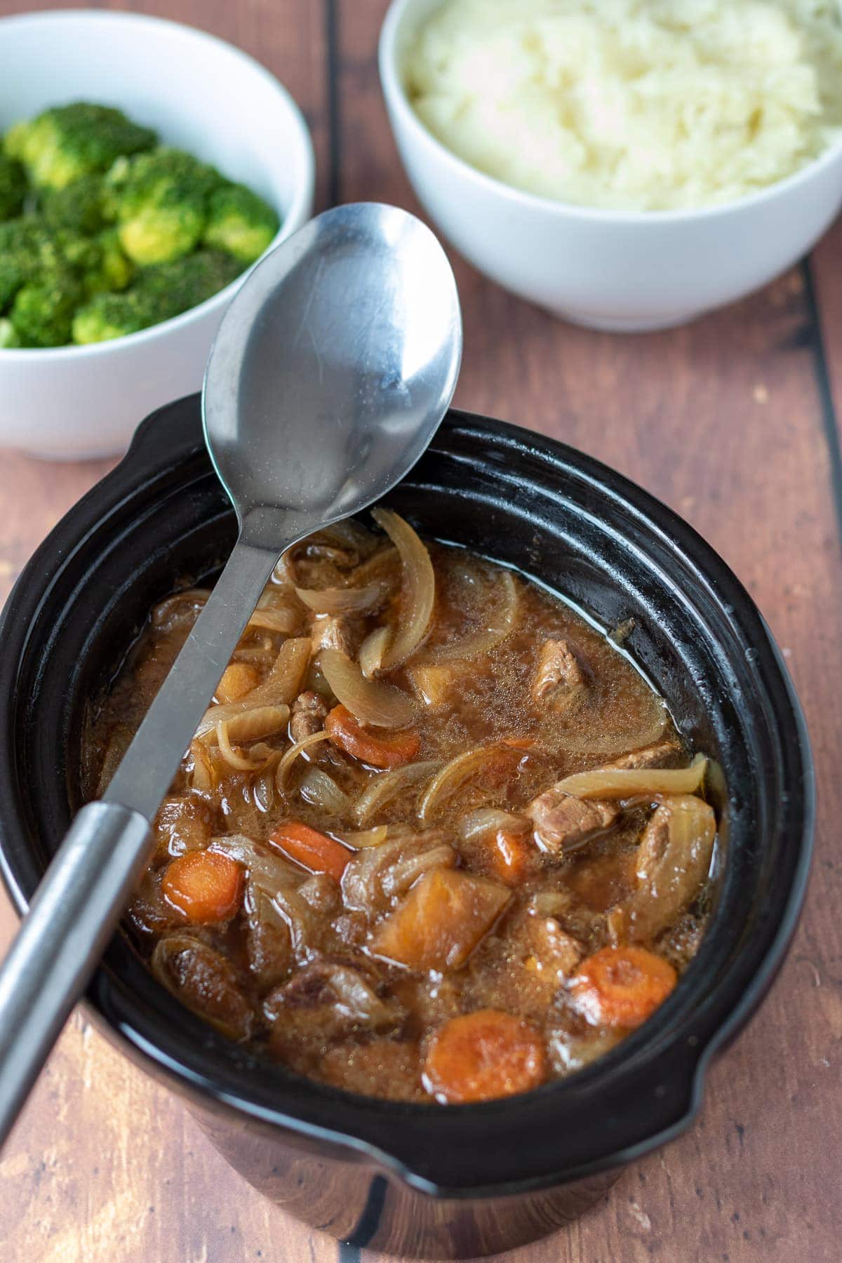 Slow cooker beef and ale stew with serving spoon, white bowl of broccoli and rice.