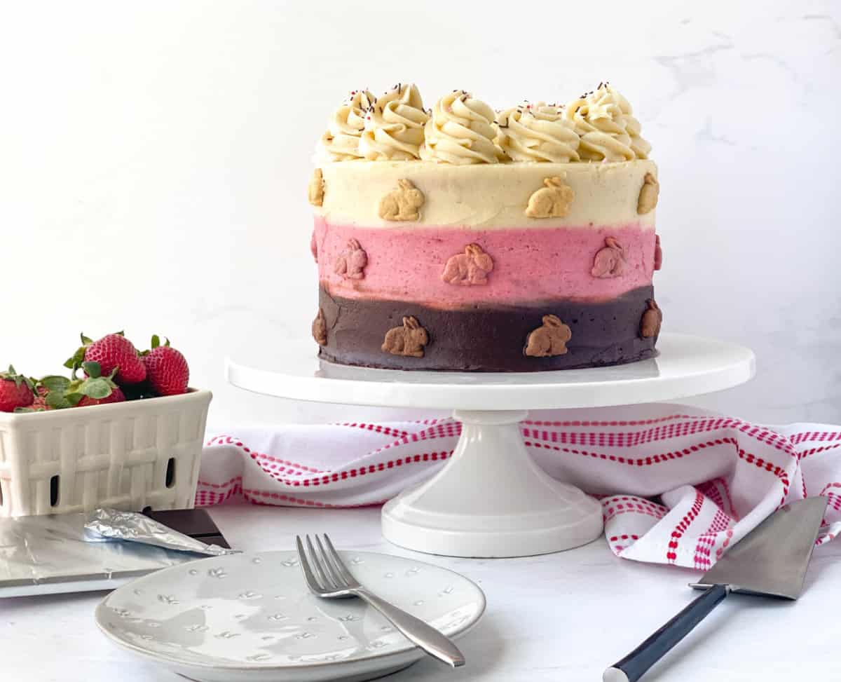 Decorative Neapolitan cake on a stand with miniature bunny grahams and a basket of strawberries.