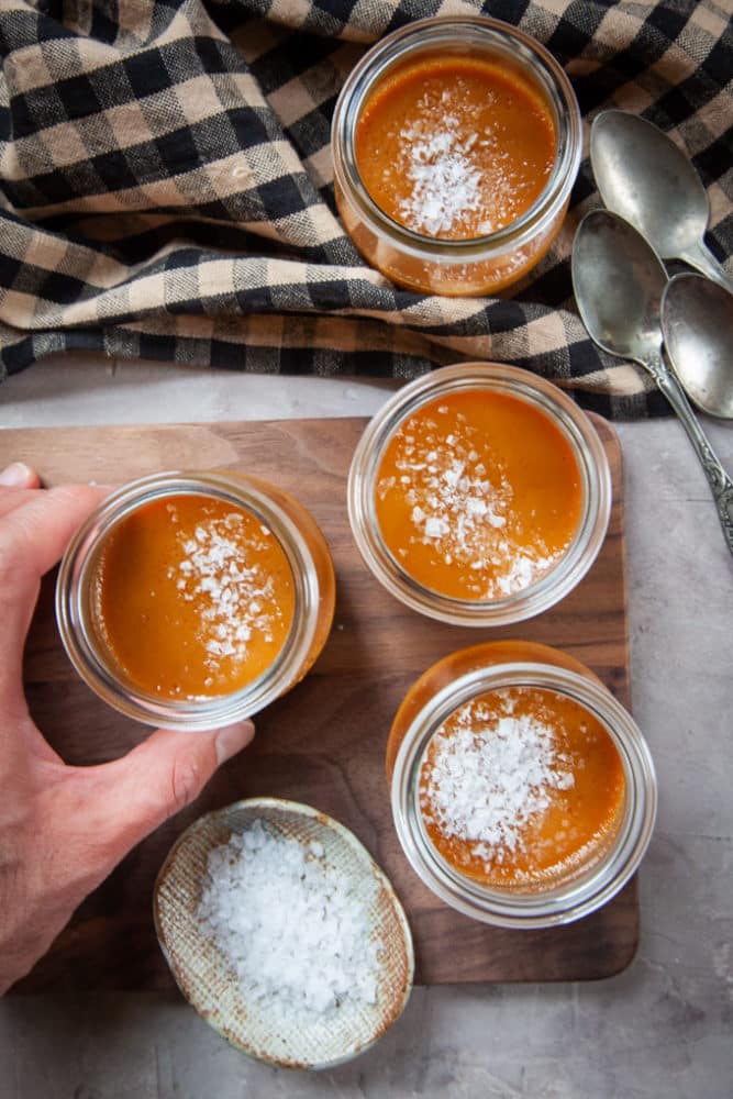 Hand holding salted caramel pot de creme with more on a cutting board.