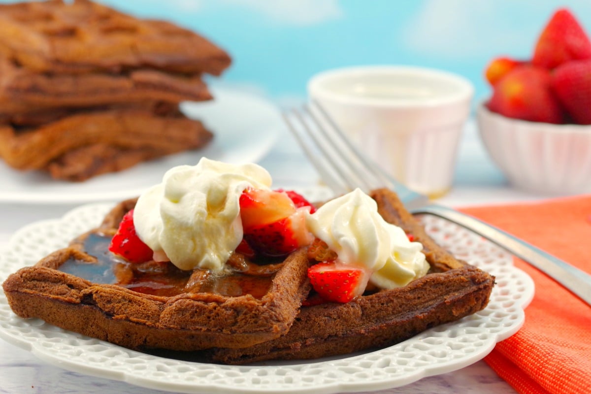 Chocolate waffles with whipped cream and sliced strawberries on a decorative plate.