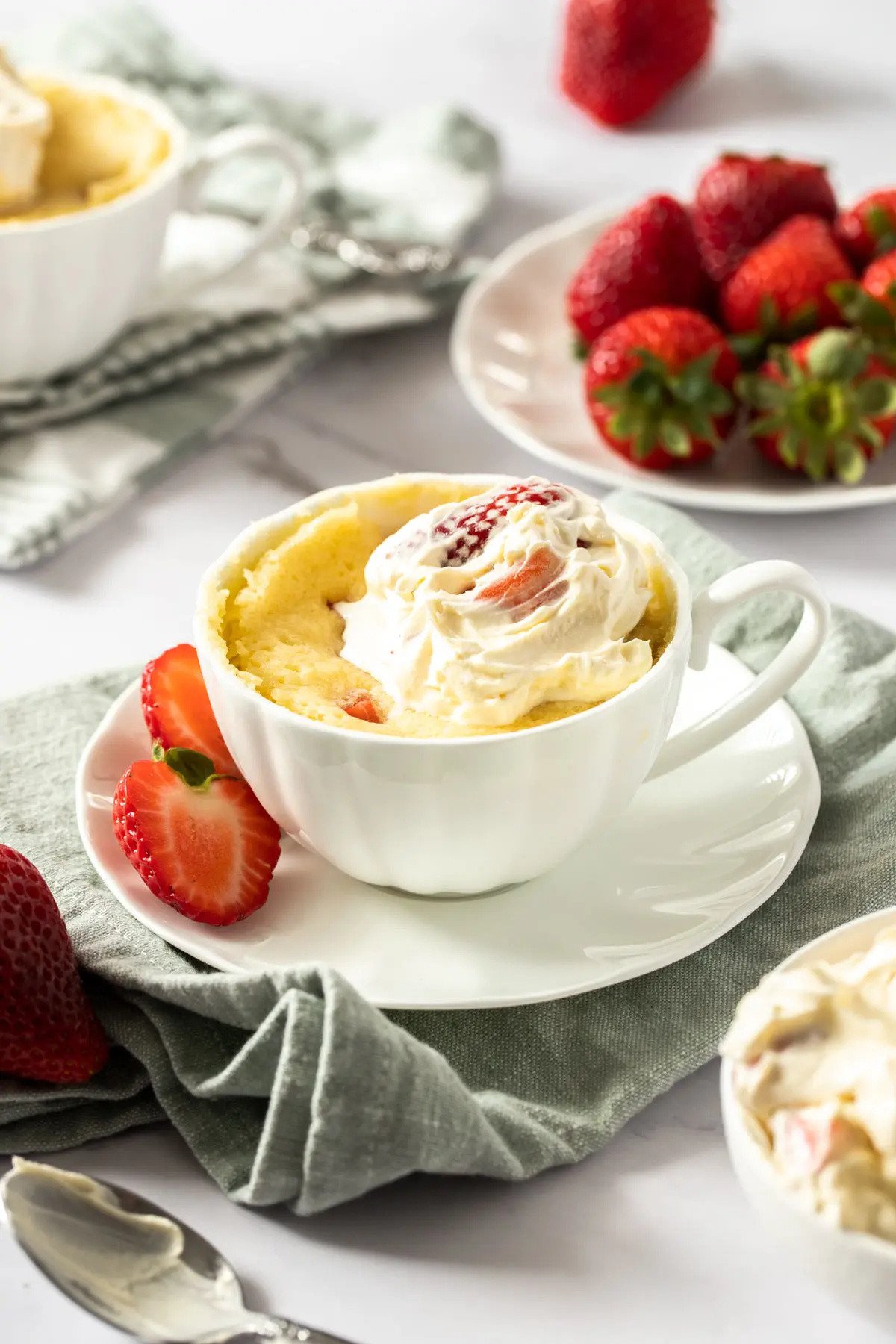 Strawberry shortcake mug cake in a tea cup with a plate of strawberries in the background.