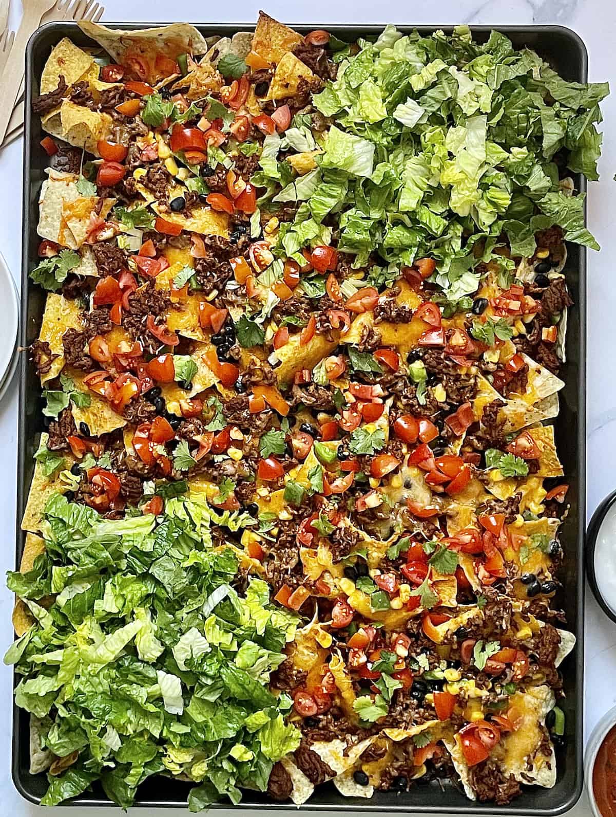 Loaded nachos with tortilla chips, ground beef, and other toppings on a sheet pan.