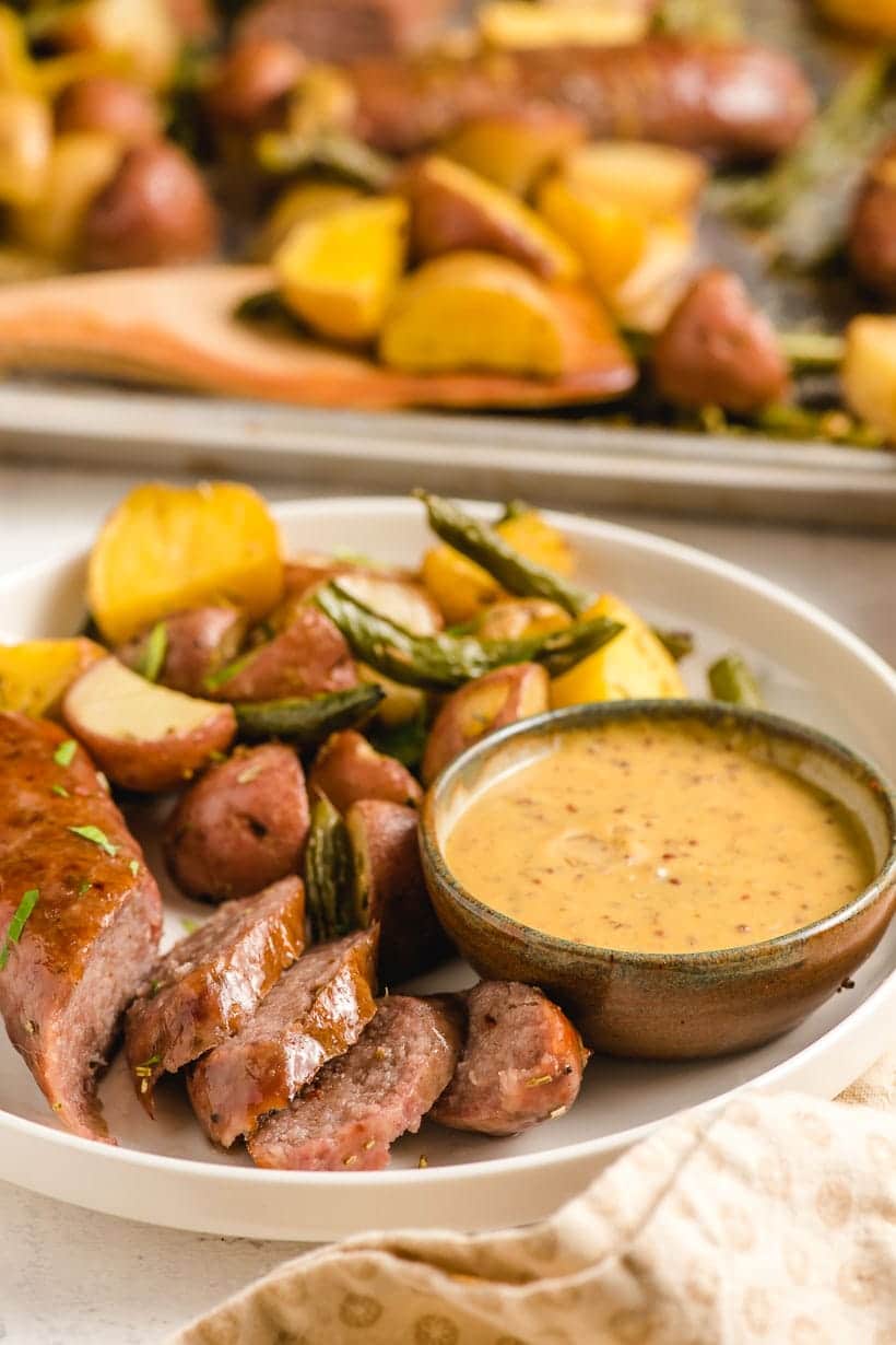 Italian sausage, green beans, and potatoes with Dijon mustard sauce on a white plate with more in the background.