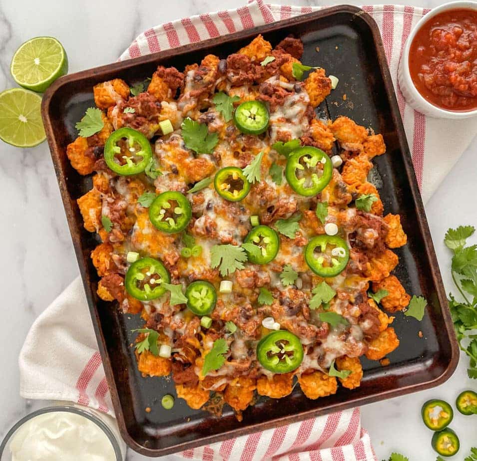 Chili cheese tater tots nachos in a pan with ingredients around it.