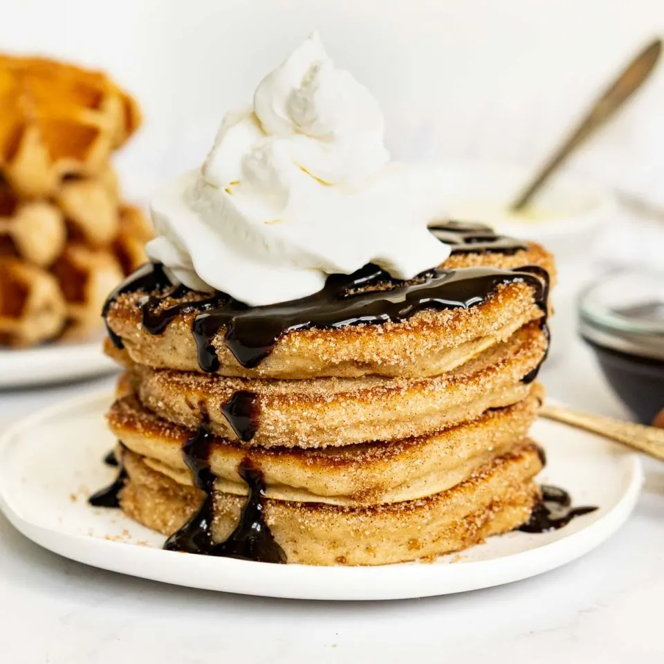 Stack of churro pancakes with chocolate sauce and whipped cream on top.