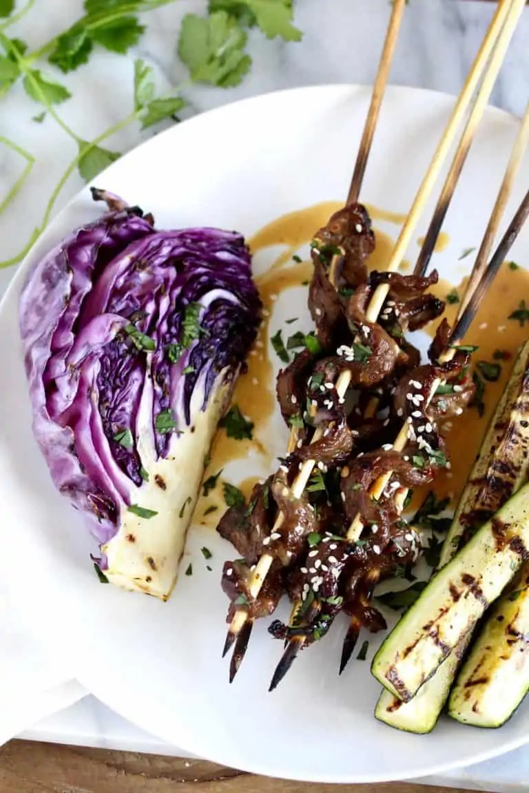 Beef satay skewers with grilled zucchini and cabbage on a white plate.