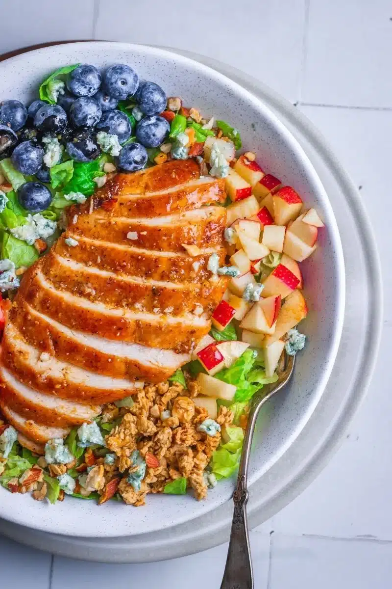 Fruit salad with sliced chicken, diced apple, blueberries, granola, and blue cheese crumbles on lettuce.