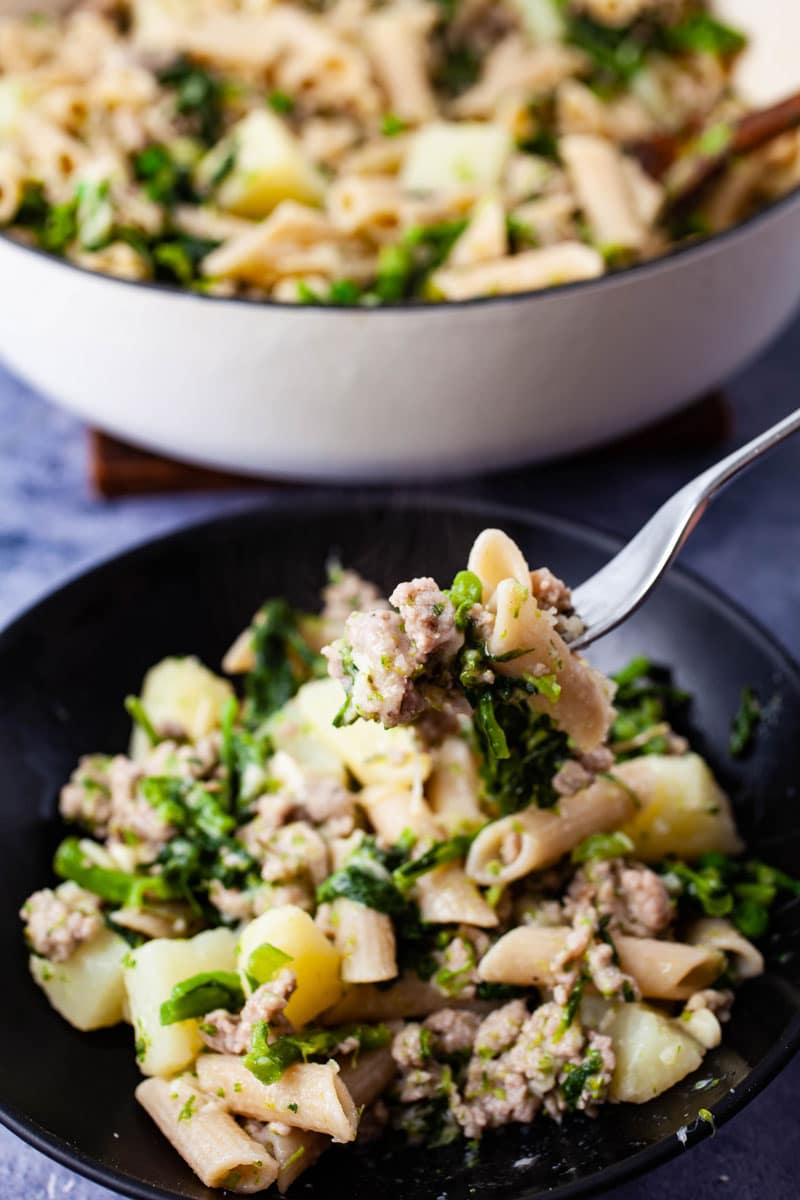 Fork with bite of sausage broccoli rabe pasta with potatoes on a black plate.