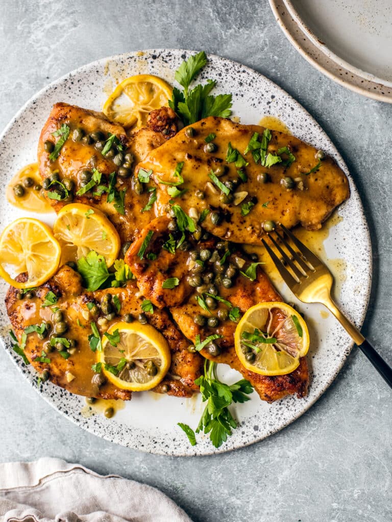 Chicken piccata with sauce, capers, and lemon on a decorative plate with a golden fork.