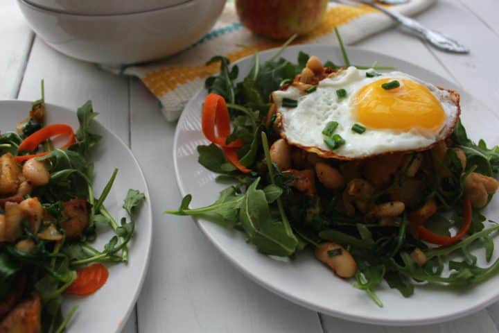 Two servings of crispy hash with fried eggs with arugula, beans, diced apple, and other ingredients with an egg on top.