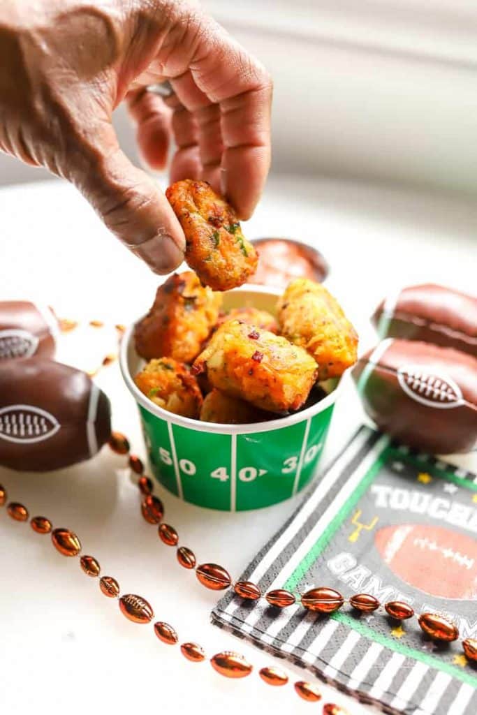 Hand holding a loaded chicken tater tot with football decorations in the background.