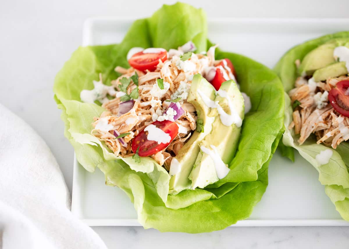 Buffalo chicken lettuce wraps with dressing, avocado, red onions, and tomatoes on a white platter.