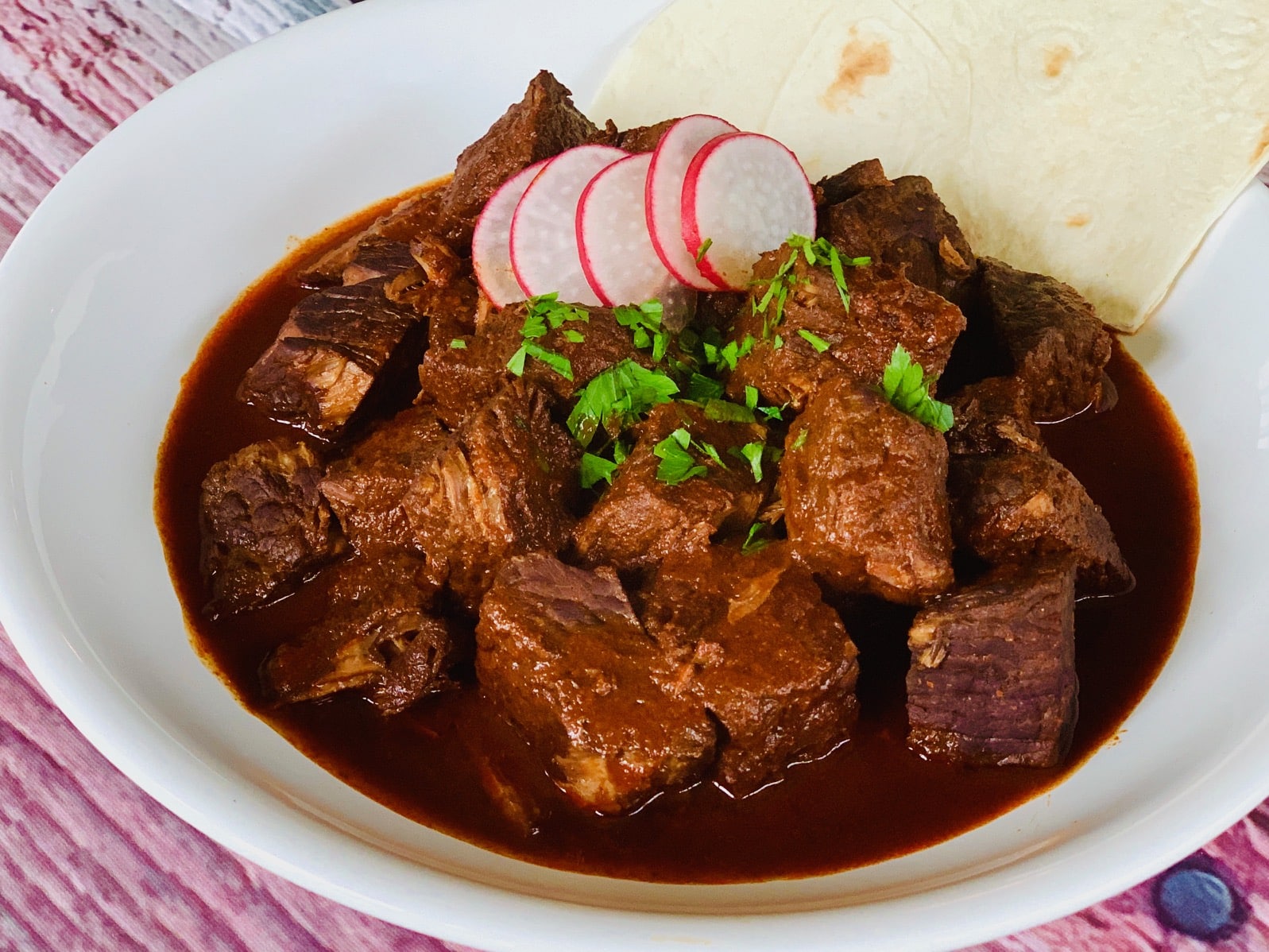 Chile Colorado with tortilla, radish, and cilantro in a bowl.