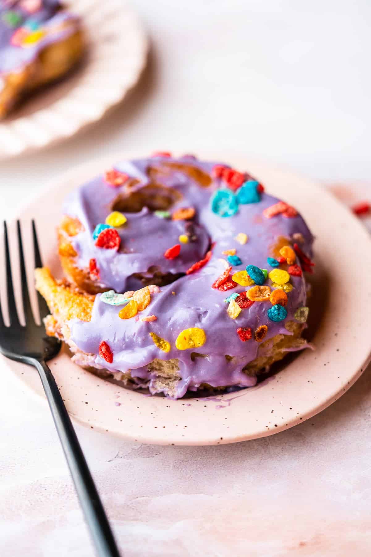 Cereal milk cinnamon rolls topped with purple ube cream cheese frosting and colorful cereal on a saucer with a fork.