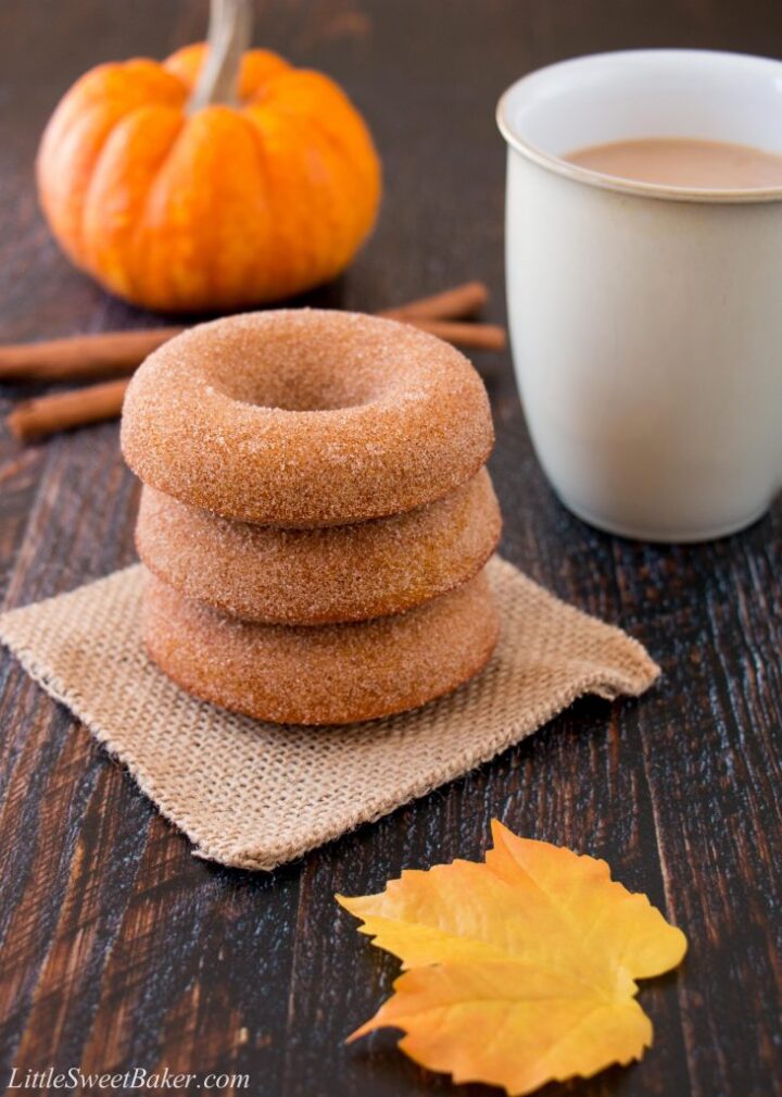 Stack of homemade pumpkin spice donuts with leaf and cup filled with coffee in the background.