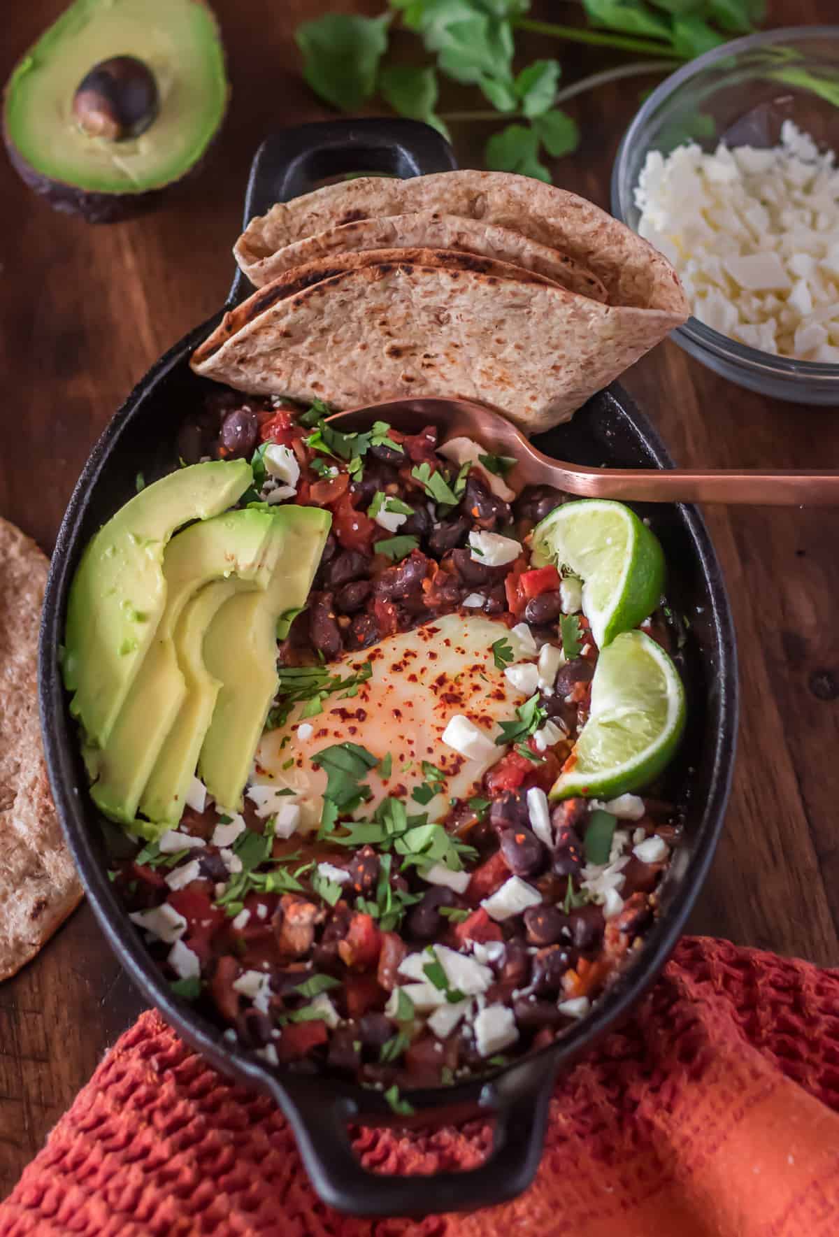 Mexican black bean shakshuka with beans, cheese, avocado, limes and tortillas.