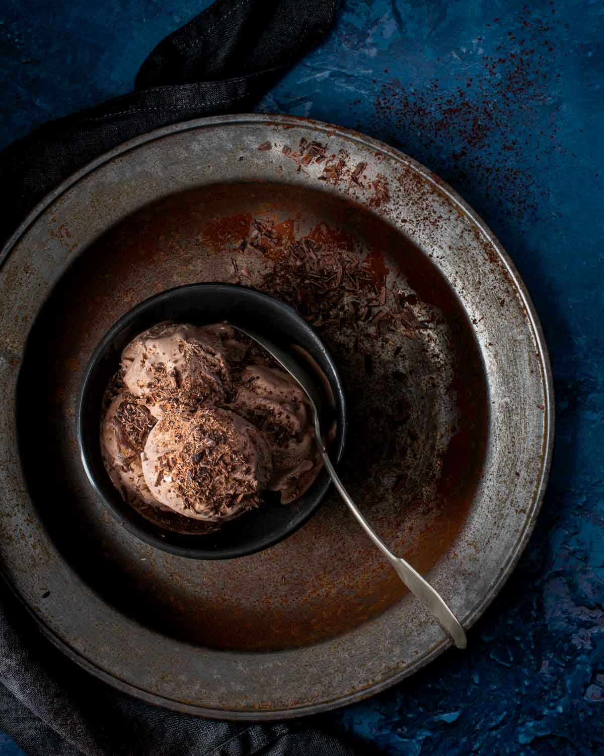 Chocolate ice cream with spoon in a bowl on a large metal plate.