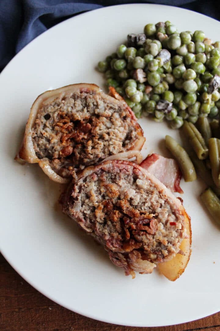 Serving of smoked bacon wrapped pork  with peas and green beans on the side on white plate.