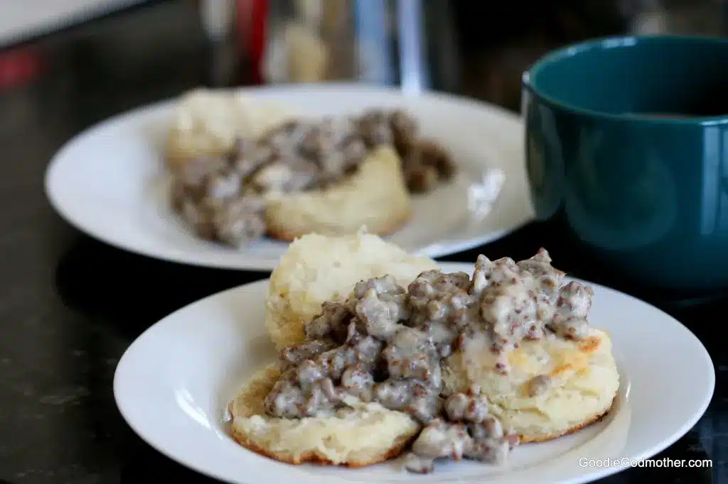 Meat lovers biscuits and gravy on a white plate with blue cup in the background.