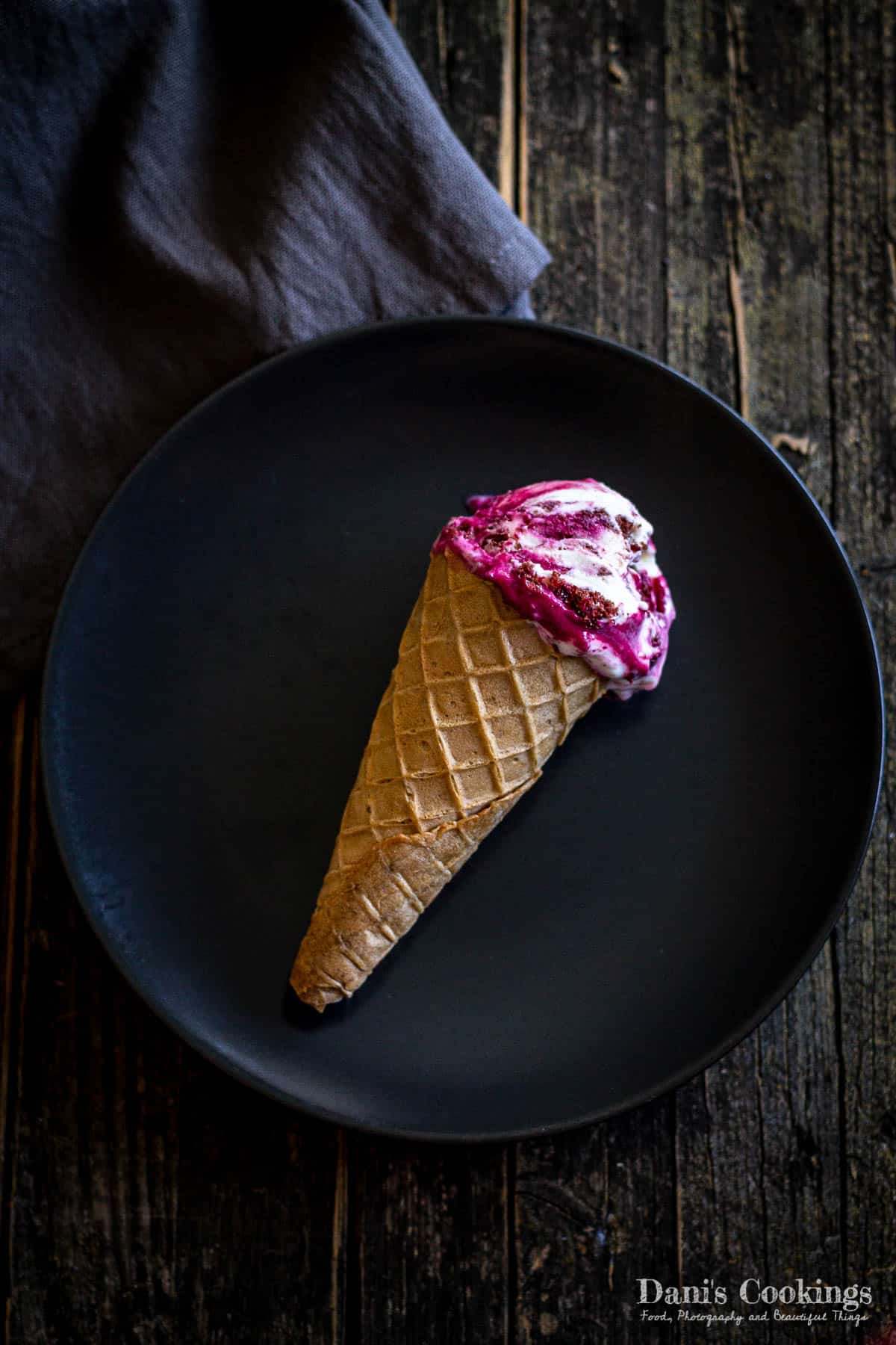 Red velvet ice cream cone on a black plate with napkin on a wooden board.